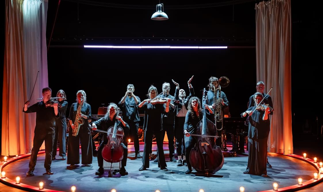 A group of musicians wearing black, standing onstage with their instruments, lit by circus bulb lights.