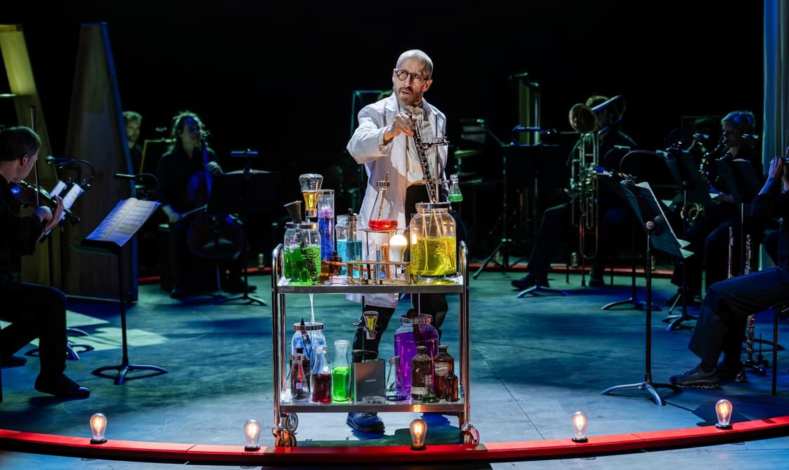 Man onstage wearing a lab coat with an trolley filled with bottles and vials of coloured lquids.