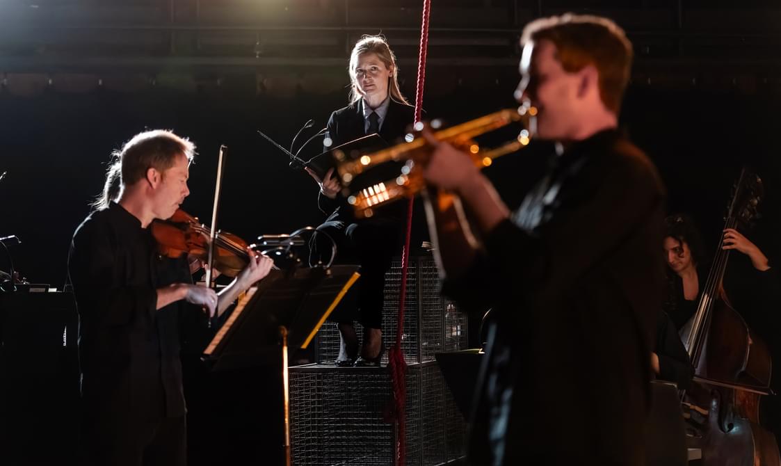 A violinist, a trumpet player and a woman holding a book arranged on stage.