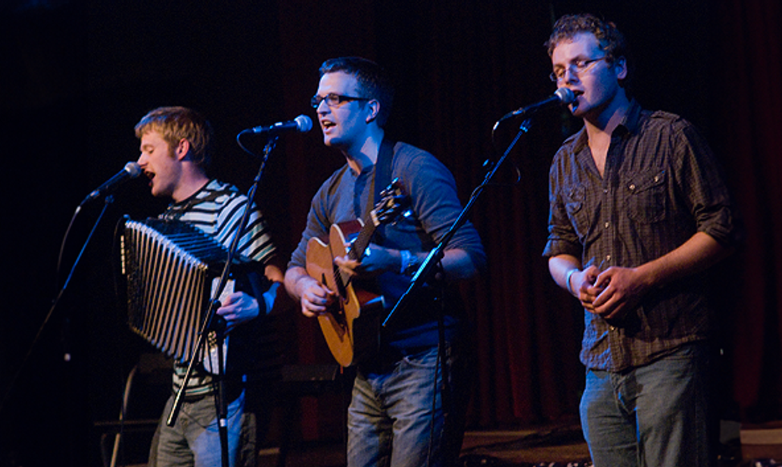 Three men standing onstage playing accordion and guitar whilst singing.