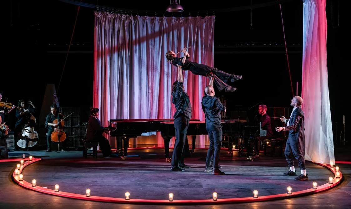 Musicians onstage, two men are holding a woman in the air as she plays the flute.