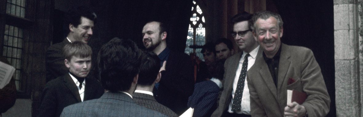 Benjamin Britten standing and smiling in the entrance of a church with a group of people standing around.