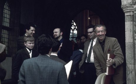 Benjamin Britten standing and smiling in the entrance of a church with a group of people standing around.
