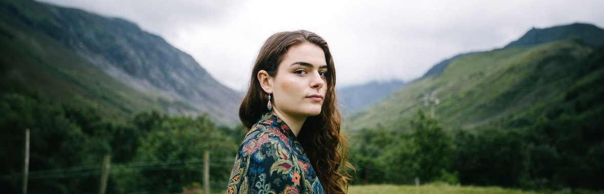 Brighde Chaimbeul, woman standingside-on and looking into the camera, against a background of green fields and mountains.