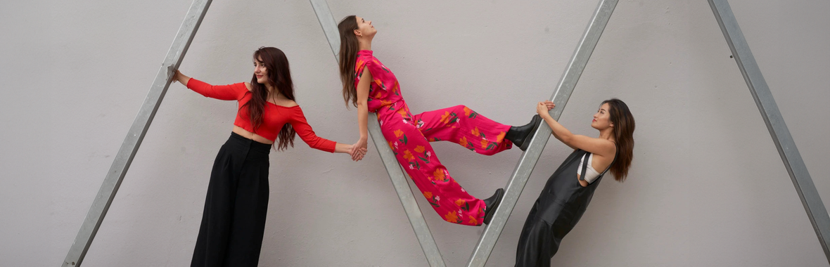 Three women from De Beauvoir Trio standing wearing pink and black, standing posed around a wooden structure.