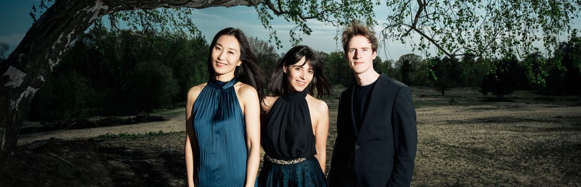 Quantum Clarinet Trio, two women and a man dressed in black and blue, smiling at the camera with trees and grass behind them.