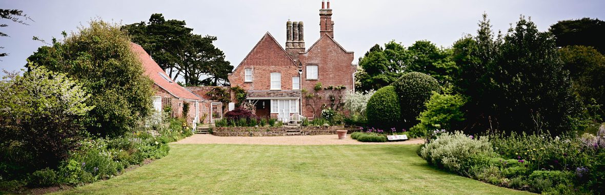 The Red House and Garden taken at a distance, red brick building surrounded by a green lawn with trees and flowers.
