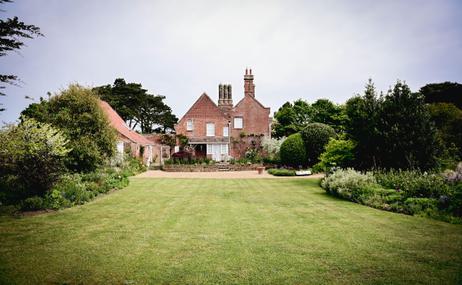 The Red House and Garden taken at a distance, red brick building surrounded by a green lawn with trees and flowers.