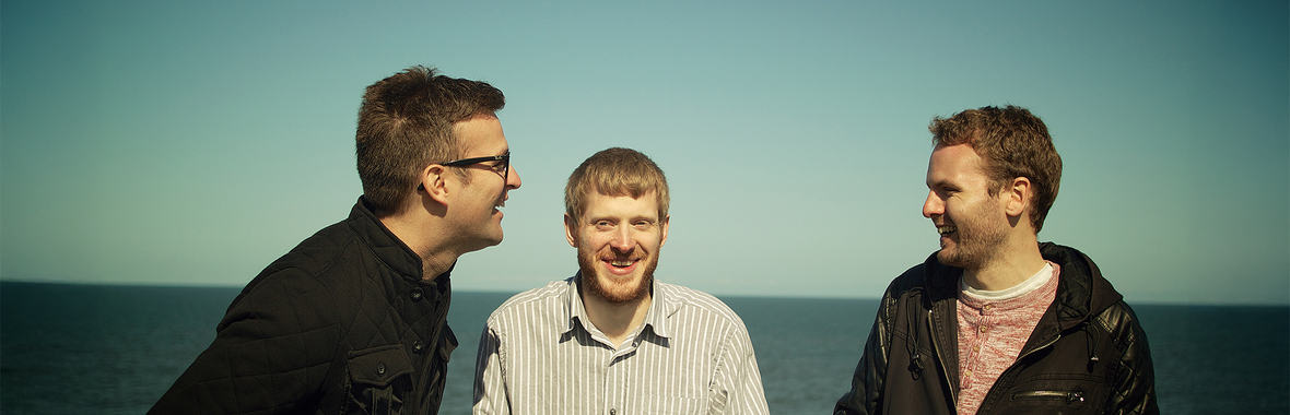Three men standing together smiling and laughing with the ocean in the background