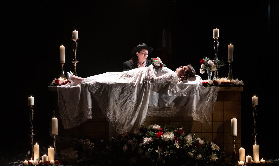 An actress in a white dress is lying on a casket holding flowers, surrounded by candles. Another actor stands beside her singing.