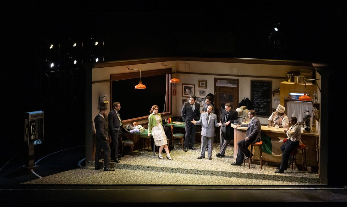 Stage setting of a pub interior, with actors and singers standing around a bar.