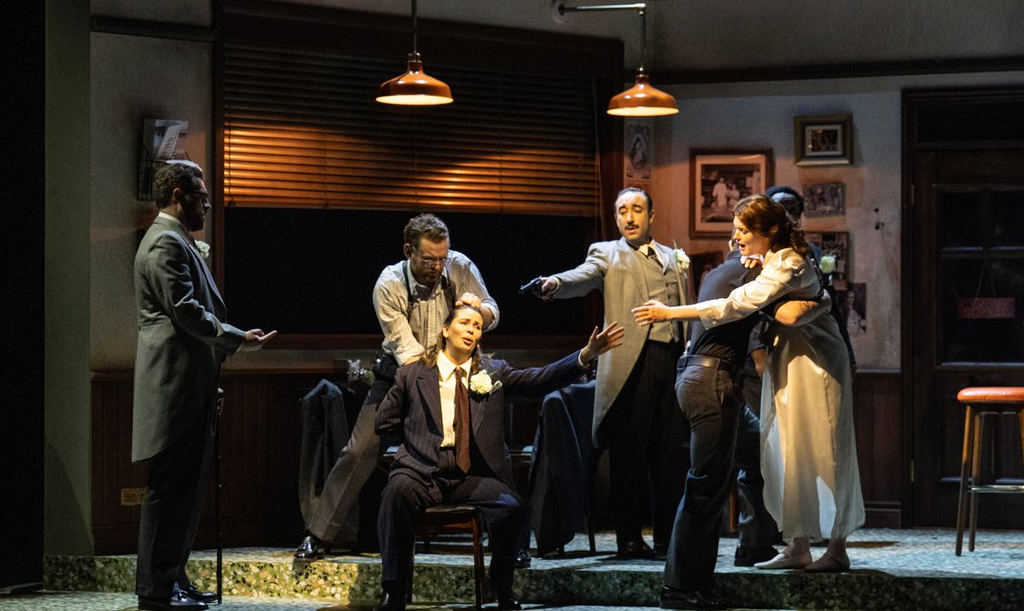 Stage setting of a pub interior, a singer is seated whilst around him standing figures are pointing guns at her.