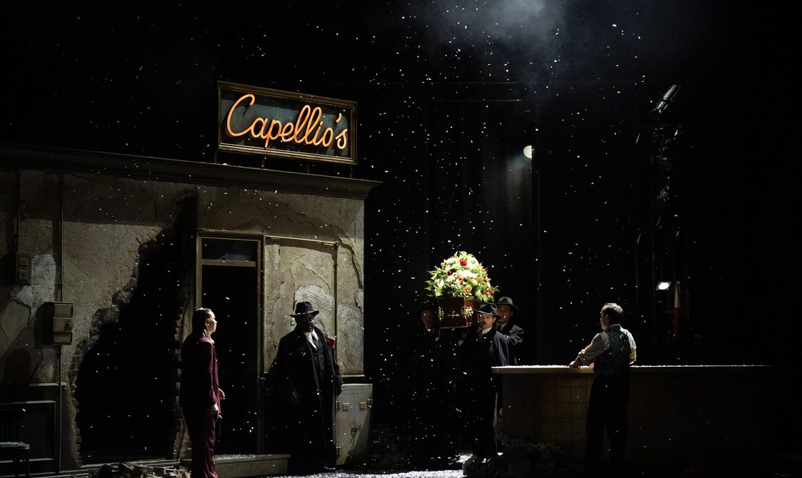 Darkly lit stage setting of a pub exterior called Capello's, silhouetted figures are entering the scene carrying a casket covered with flowers.