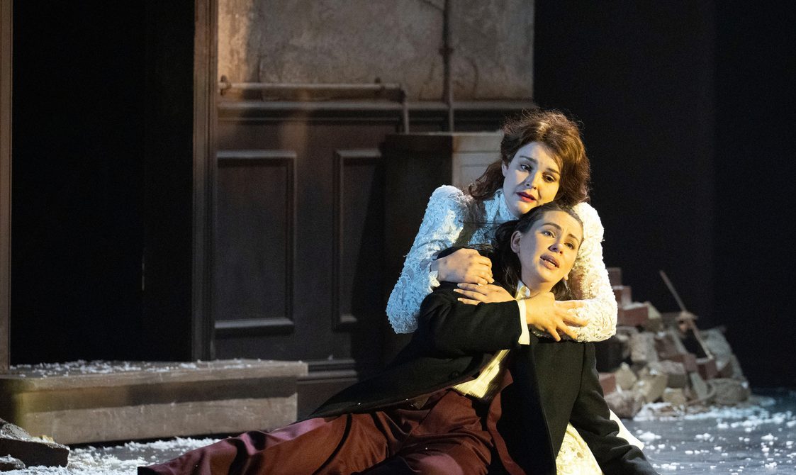 Two singers dressed in period lying down and embracing each other.