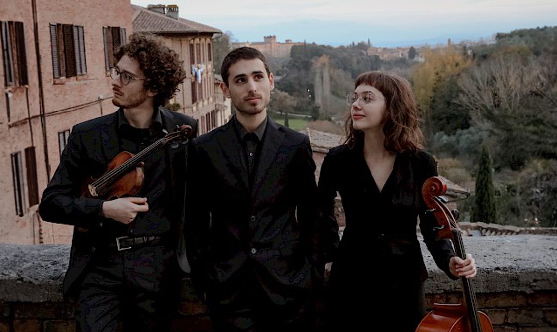 Trio Rinaldo, two men and a woman standing outside holding their string instruments with buildings and trees behind them.