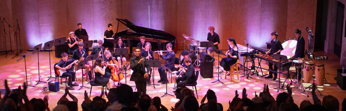 Aldeburgh Young Musicians standing and sitting onstage, playing their instruments.