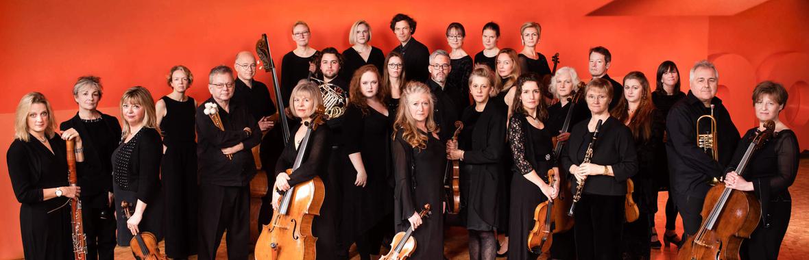 The Britten Sinfonia holding various instruments standing against a orange wall.