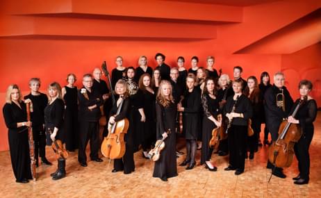 The Britten Sinfonia holding various instruments standing against a orange wall.