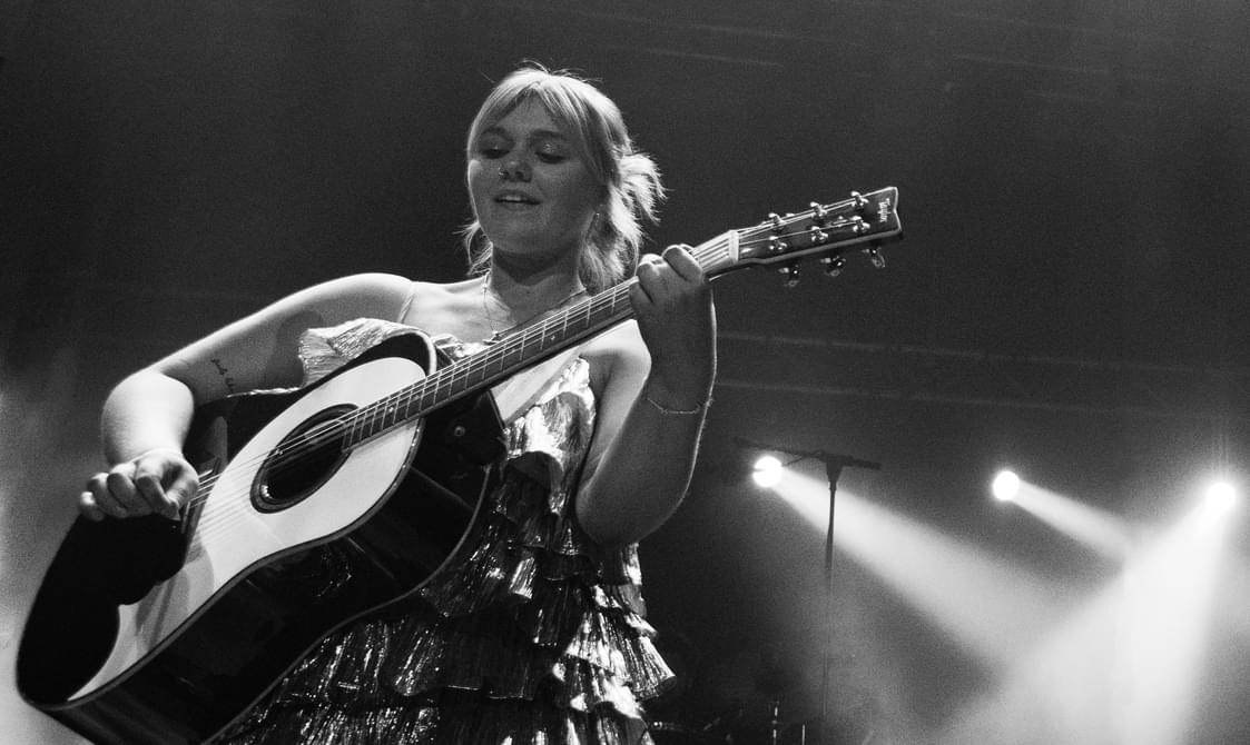 Black and white audience view of a young woman with blond tied-back hair wearing a frilly dress and playing the guitar.