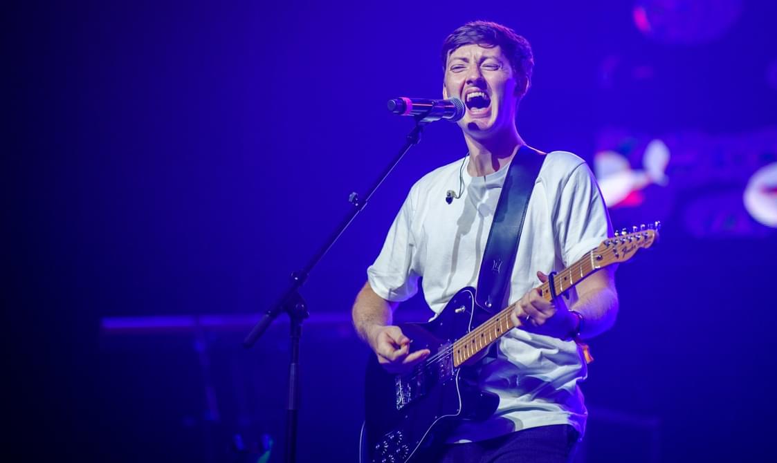 Young man with short hair in a white tshirt, playing the guitar and singing into a microphone, bathed in soft blue light.