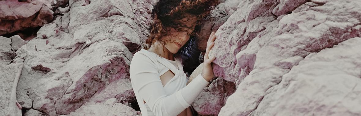 Young woman wearing white, standing amongst the rocks with brown hair blowing in the wind.