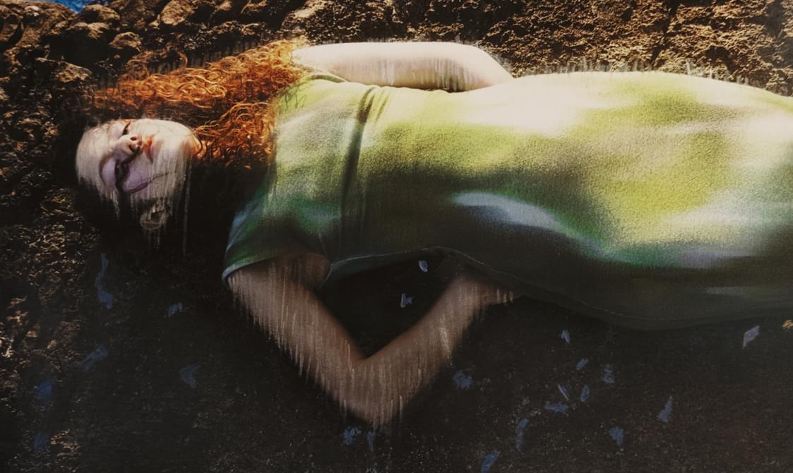 Woman with long curly hair wearing a green dress and lying submerged in water with her eyes closed.
