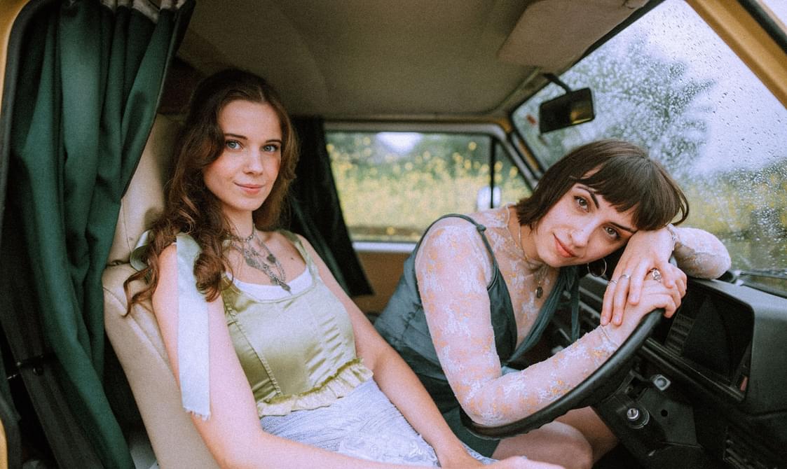 Two young women sitting in the front seats of a car and looking at the camera, one is sitting back in the seat while the other has her arms draped over the steering wheel.