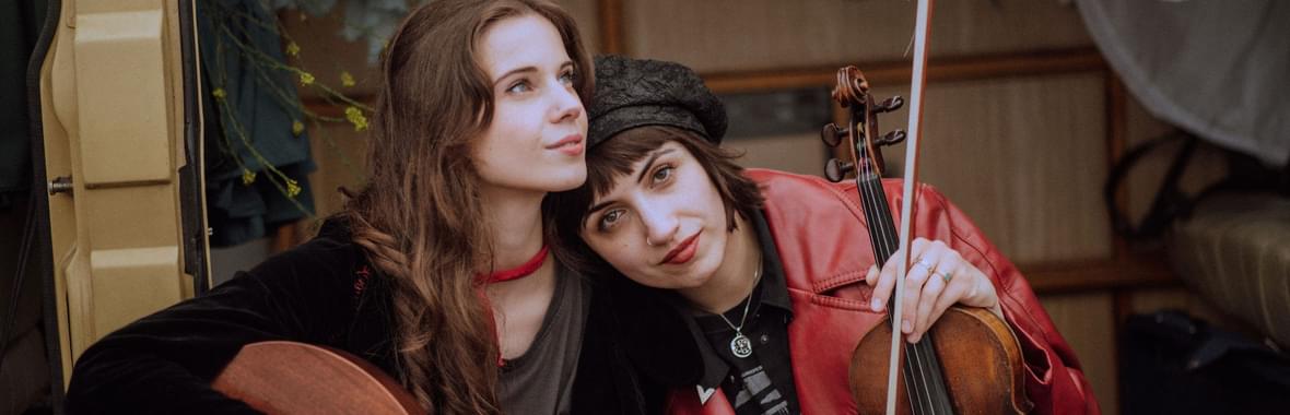 Two young women sitting close together outside, one with a guitar and the other with a violin.