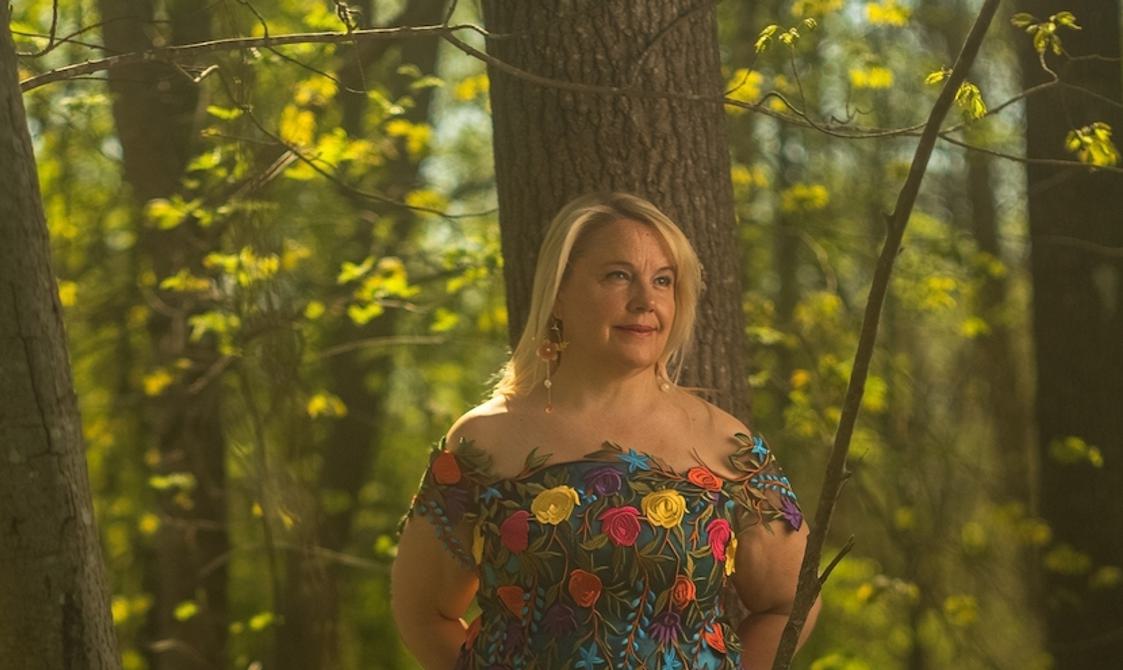 Blond woman in a floral dress leaning against a tree in a forest.