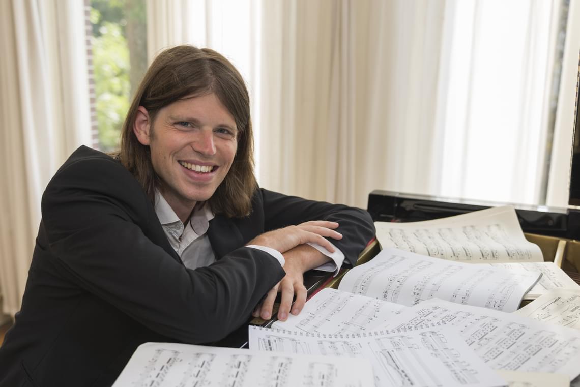 Man with long brown hair sitting at a piano with music sheets and smiling.