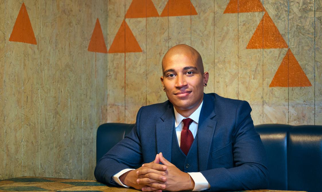 Daniel Kidane, wearing a blue suit, sitting at a table and smiling at the camera.