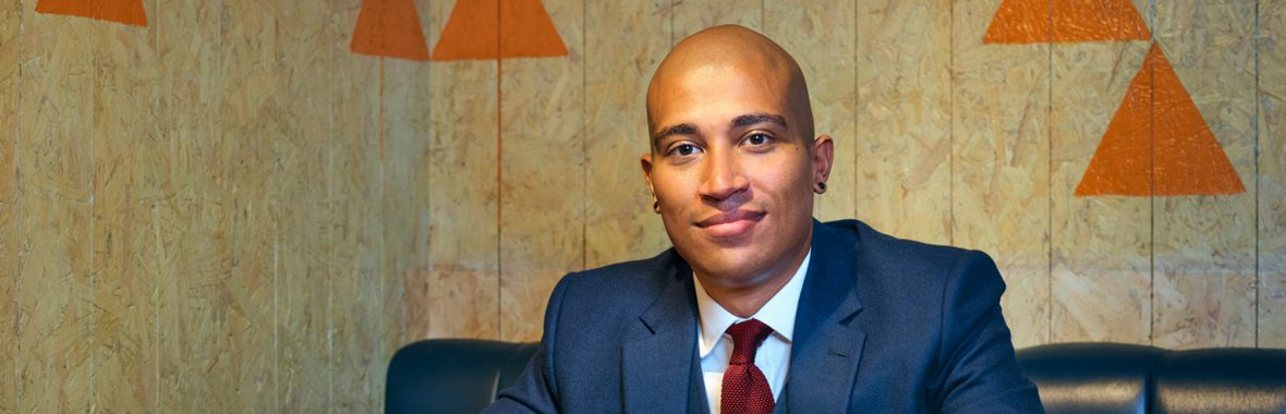 Daniel Kidane, wearing a blue suit, sitting at a table and smiling at the camera.