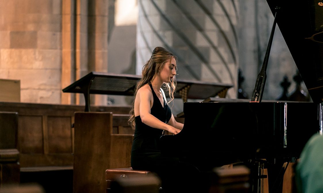 Francesca Lauri, young woman in a sleeveless black dress, seated at and playing a grand piano.