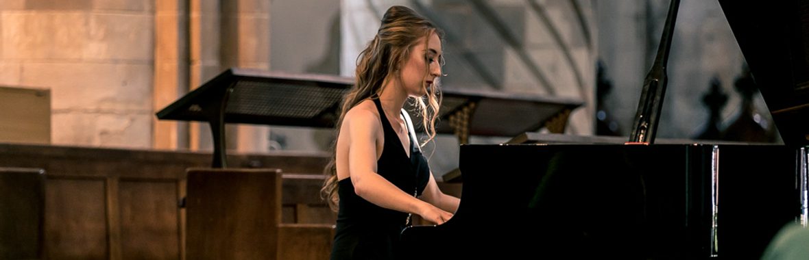 Francesca Lauri, young woman in a sleeveless black dress, seated at and playing a grand piano.