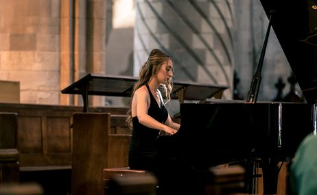 Francesca Lauri, young woman in a sleeveless black dress, seated at and playing a grand piano.