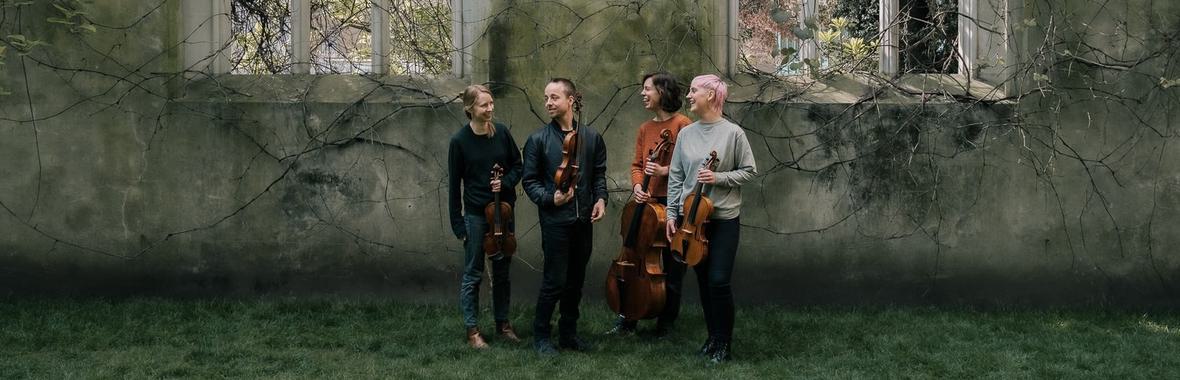 Group of four musicians holding string instruments, standing outside beneath ancient church windows