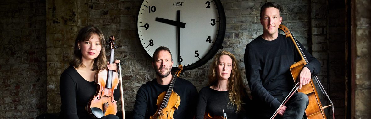 String quartet sitting under a large clock, holding their instruments.