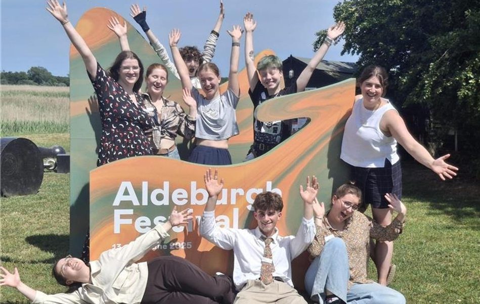 Group of young people gathered around a sign for Aldeburgh Festival 2025, holding up their arms and smiling.