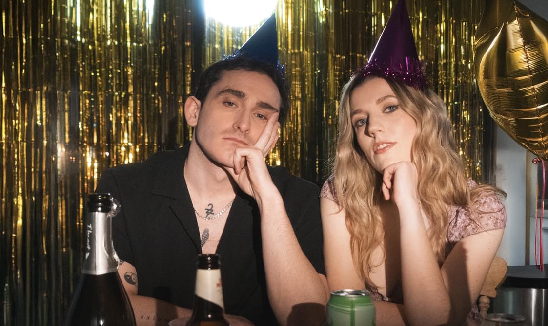 Young man with short brown hair and young woman with long blond hair wearing party hats, surrounded by glittery decorations and balloons.