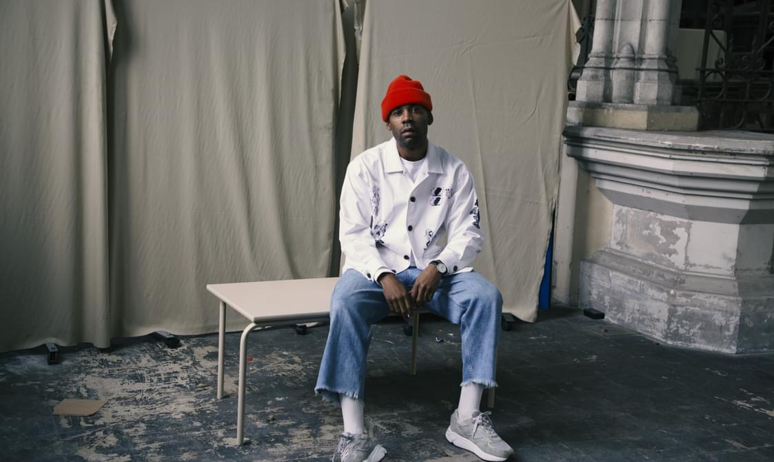 Man wearing a white jacket and red hat, sitting on a table with sheets hung up behind him.