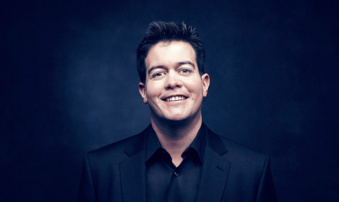 Man with short dark hair wearing a dark jacket and smiling into the camera.