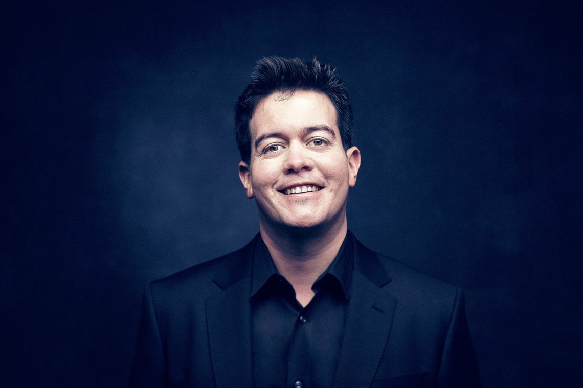 Man with short dark hair wearing a dark jacket and smiling into the camera.