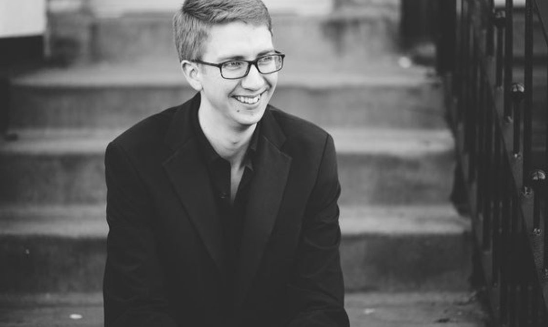 black and white image of a man with short hair and glasses, sitting on a set of steps and smiling.