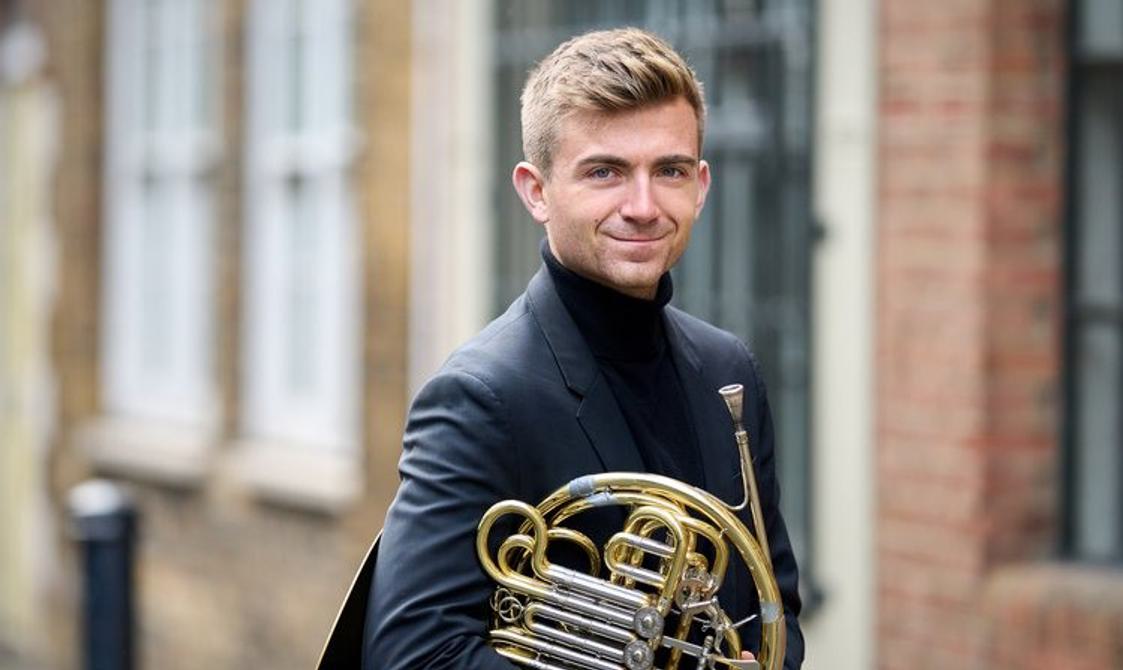 Blond man holding a french horn and smiling.