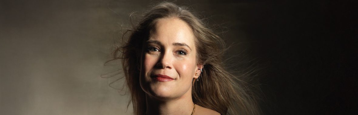 Woman with long brown hair wearing a necklace and looking into the camera, against a dark background.