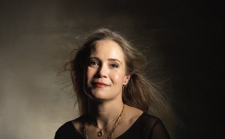 Woman with long brown hair wearing a necklace and looking into the camera, against a dark background.
