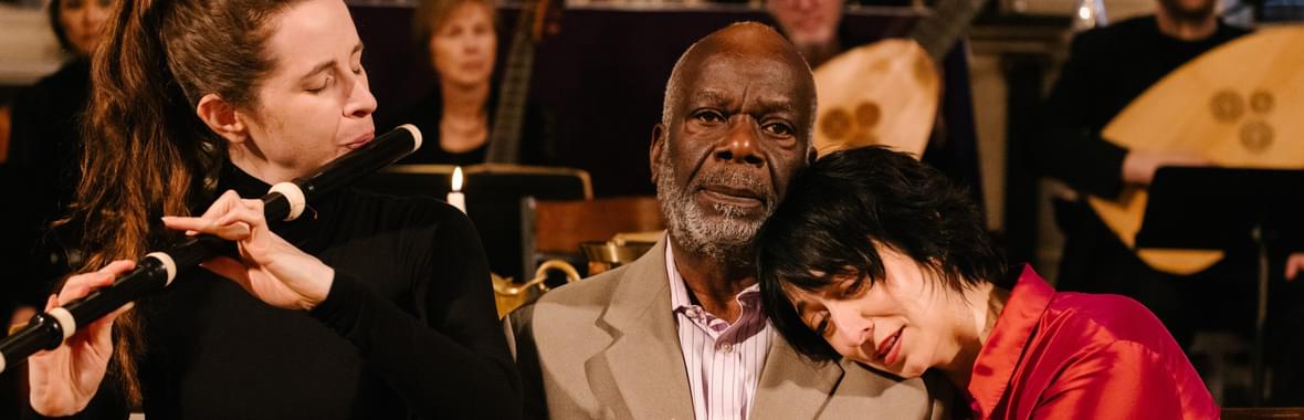 Woman playing a wind instrument sitting beside a man in a suit whilst another woman rests her head on his shoulder.