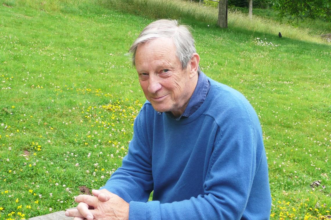 Man with short white hair wearing a blue sweater, sitting outside with grass and flowers in the background.