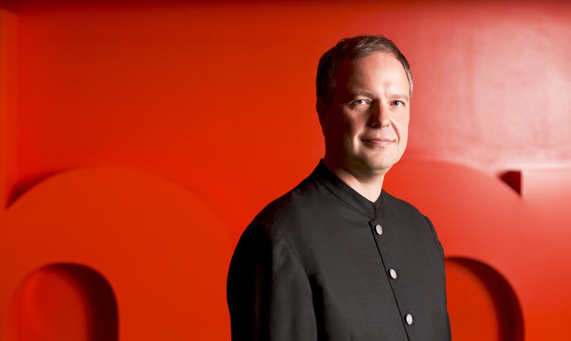 Conductor Sakari Oramo, man with cropped hair standing in front of a red background