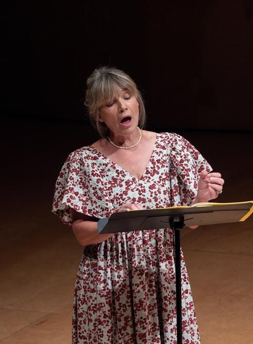 Woman wearing a floral dress, standing on stage and singing.